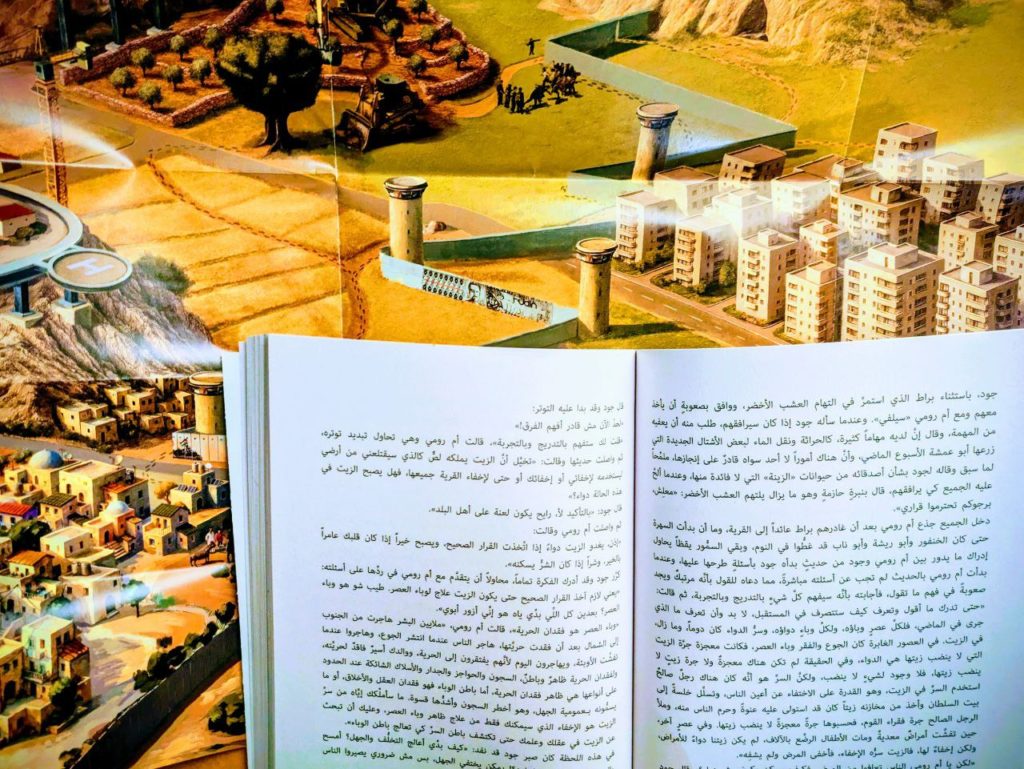 Le livre est ouvert : sur les pages, le texte est écrit en arabe.
Dans le fond, on voit une carte illustrée représentant un paysage de Cisjordanie. On voit un village palestinien, le mur de séparation et une village avec des immeubles.