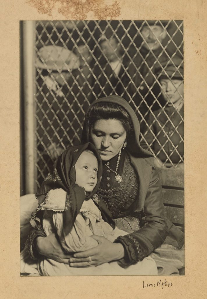 Mère très brune avec un foulard tenant son enfant contre elle. Derrière, il y a un grillage et des hommes regardent.