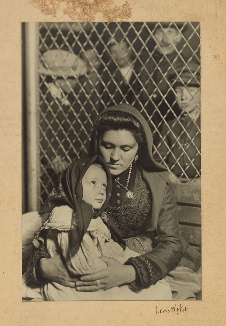 Mère très brune avec un foulard tenant son enfant contre elle. Derrière, il y a un grillage et des hommes regardent.