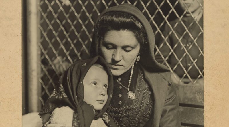 Mère très brune avec un foulard tenant son enfant contre elle. Derrière, il y a un grillage et des hommes regardent.